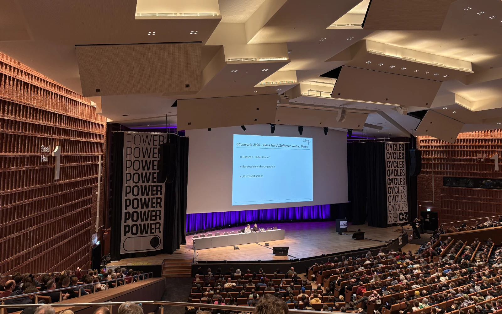 The talk about Security Nightmares: The large stage with two people sitting at a long desk with a large screen behind them and a full auditorium in front of them