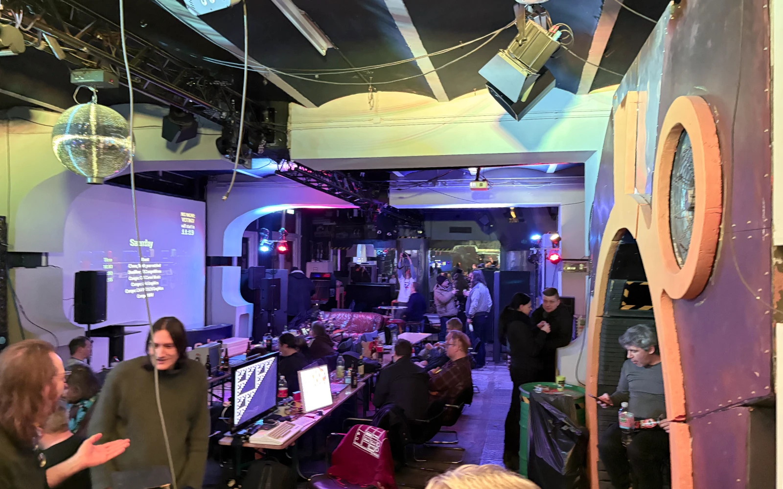 The main room of the BCC 2026 demoparty with many tables with computers on them and people talking to each other.