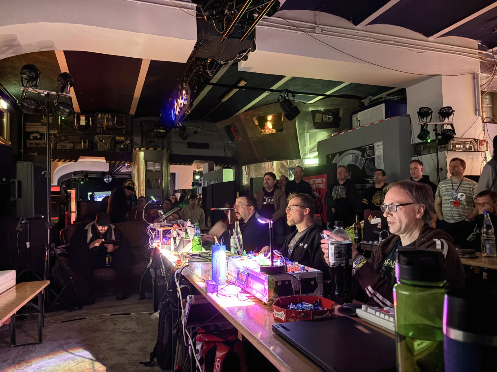 A dark room with a large projected screen showing C64 demos and a dozen or so people sitting at long tables and watching the screen.