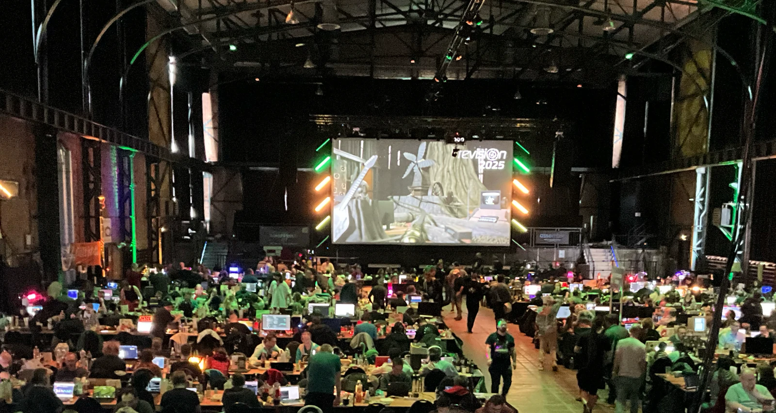 Another perspective of the main hall of the Revision 2025 demoparty, still showing table rows filled with people working on laptops but also the main stage and big screen.