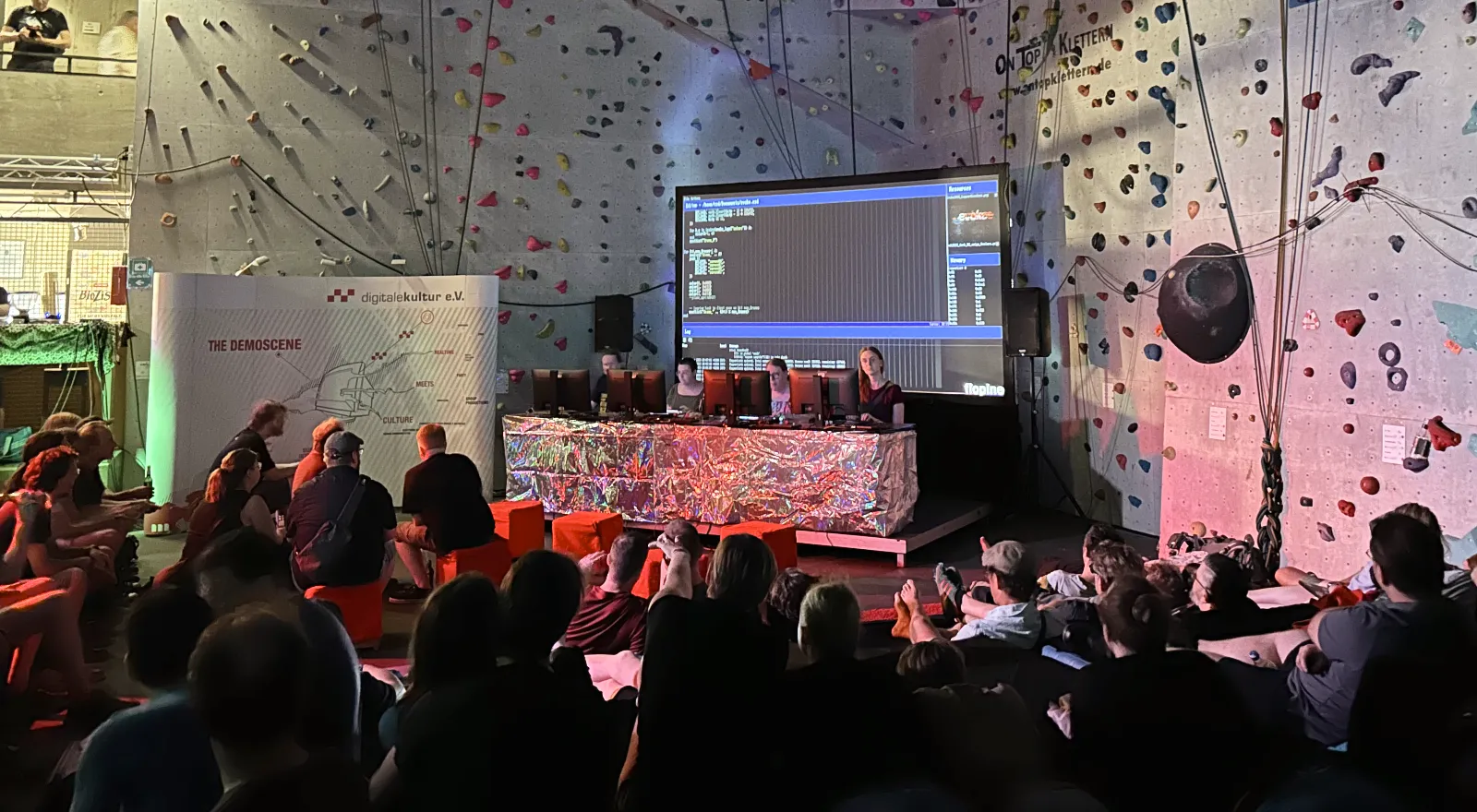 Four participants on stage with their computers and a large screen behind them showing the code they are working on.