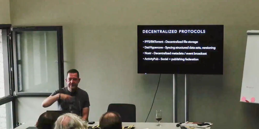 Madison, a middle-aged man, sitting at a desk before an audience presenting his slide deck on a screen behind him.