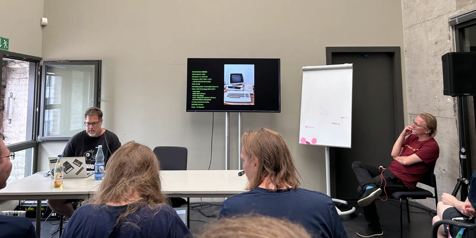 Bodo, a tall middle-aged man, sitting at a desk next to a large presentation screen in front of the audience presenting about the PET.