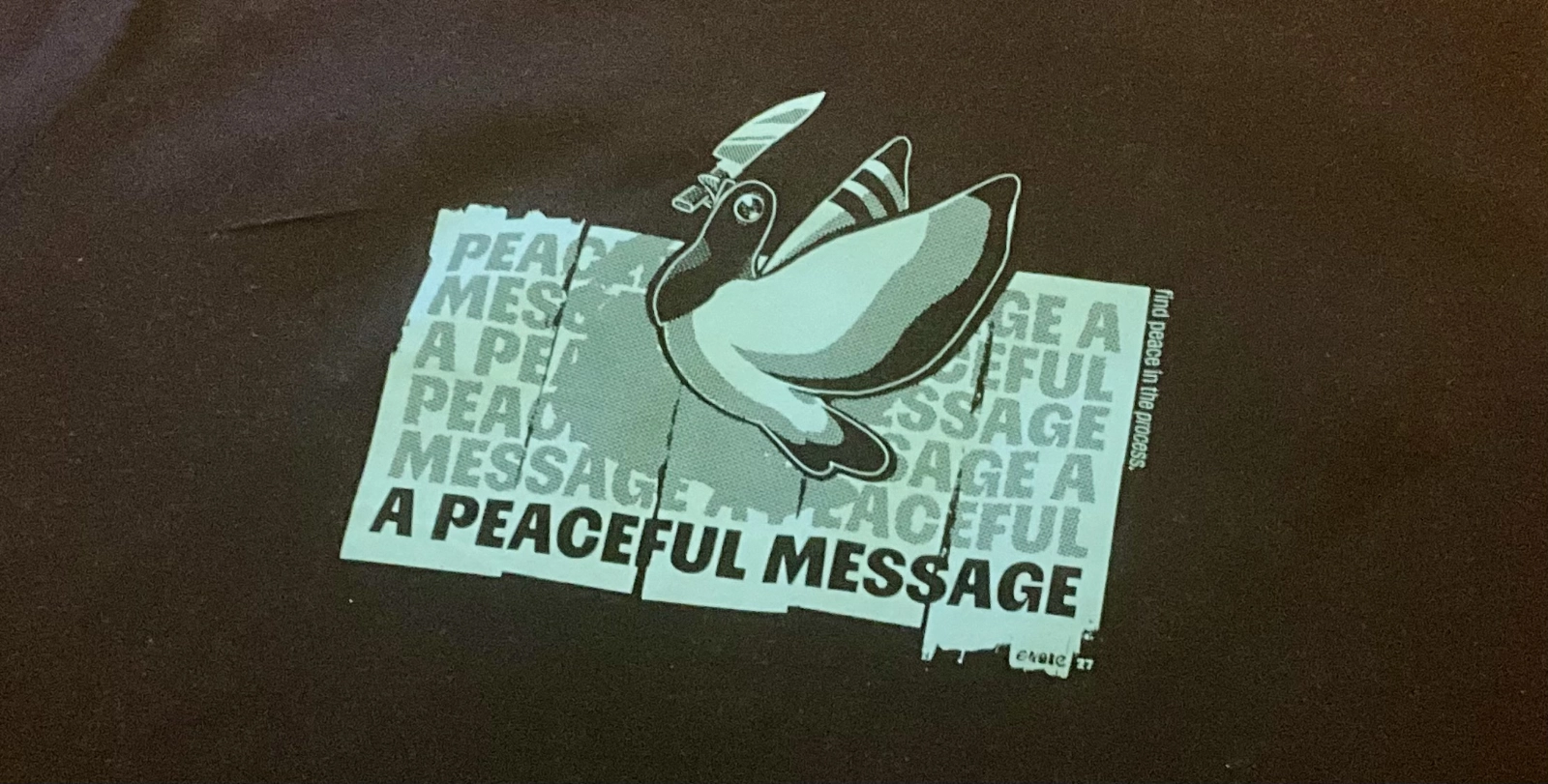The Evoke 2026 t-shirt, a dark shirt screen-printed in pale mint with a dove gripping a switchblade above tiled text reading A Peaceful Message, and find peace in the process printed down one side.