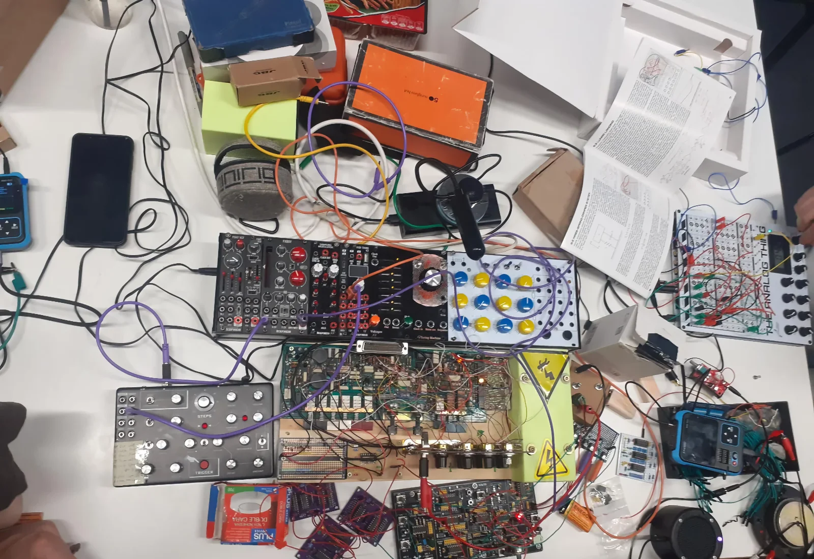 Top-down view of a workshop table covered in synthesizer modules, breadboards, patch cables, and power strips.