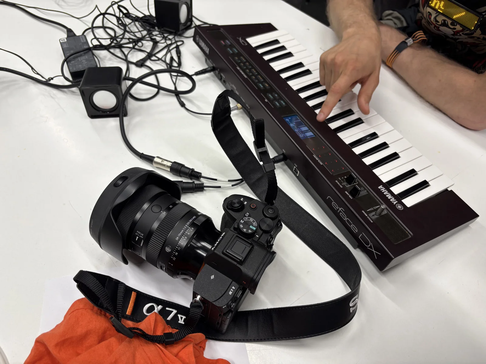 A Yamaha Reface DX FM synthesizer on a workshop table, with a hand adjusting its touch sliders and a Sony Alpha 7 IV camera in the foreground.