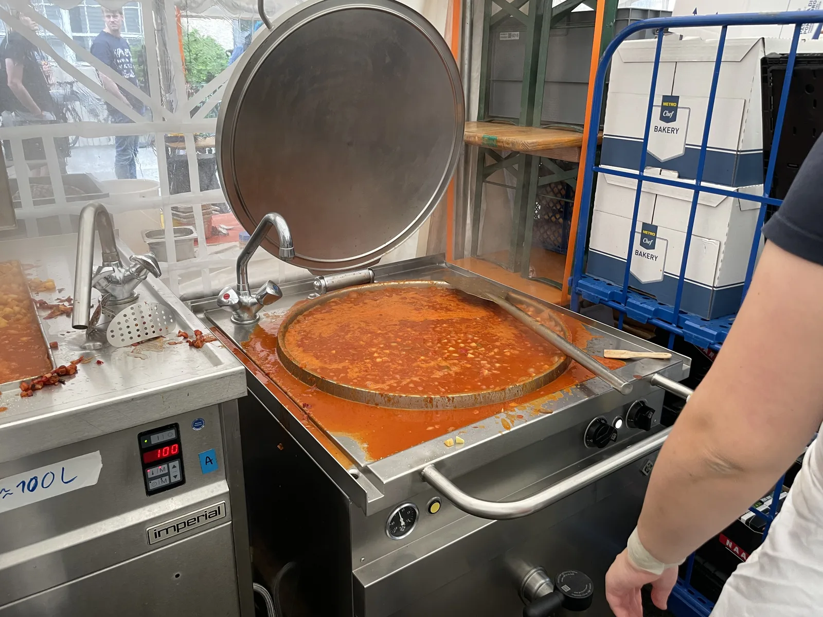 A large stainless steel tilting pan full of goulash, lid raised, in the field kitchen tent. A hand-written label on the neighboring unit reads "100 L".