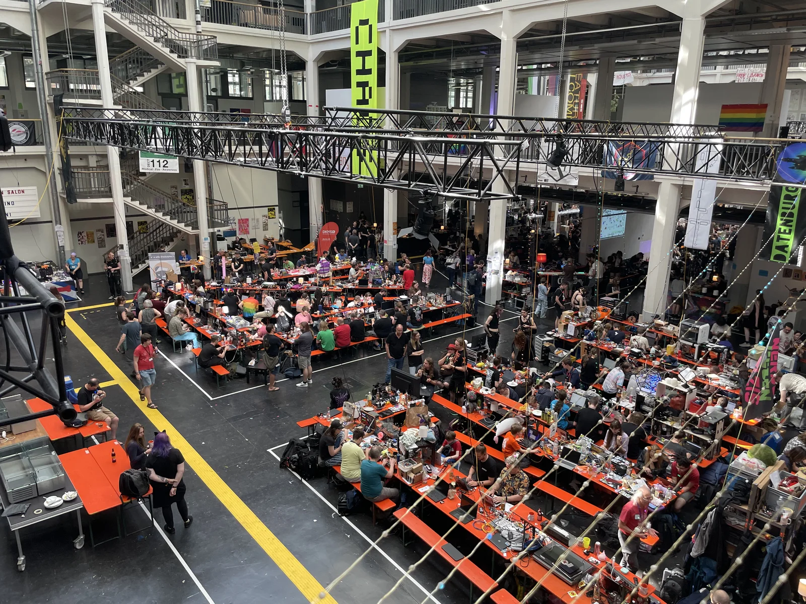View from a balcony over the large hall, rows of orange tables packed with hackers and their equipment, a green CHAOS banner and a lighting truss overhead.