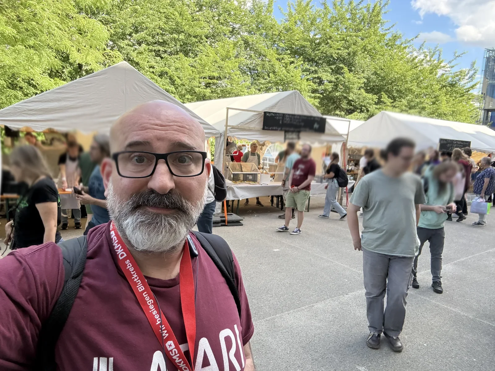 Marin standing in front of the white market tents outside the ZKM on a bright June day, with GPN24 visitors browsing the stalls behind him.