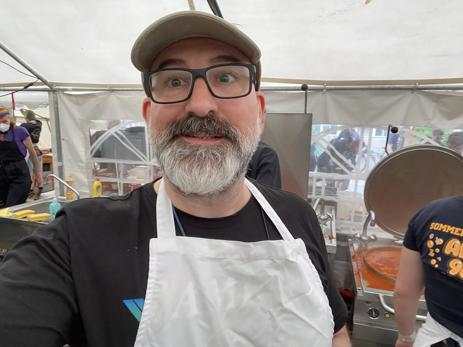 Marin in a white apron and cap in the field kitchen tent, wide-eyed, goulash machine steaming behind him.