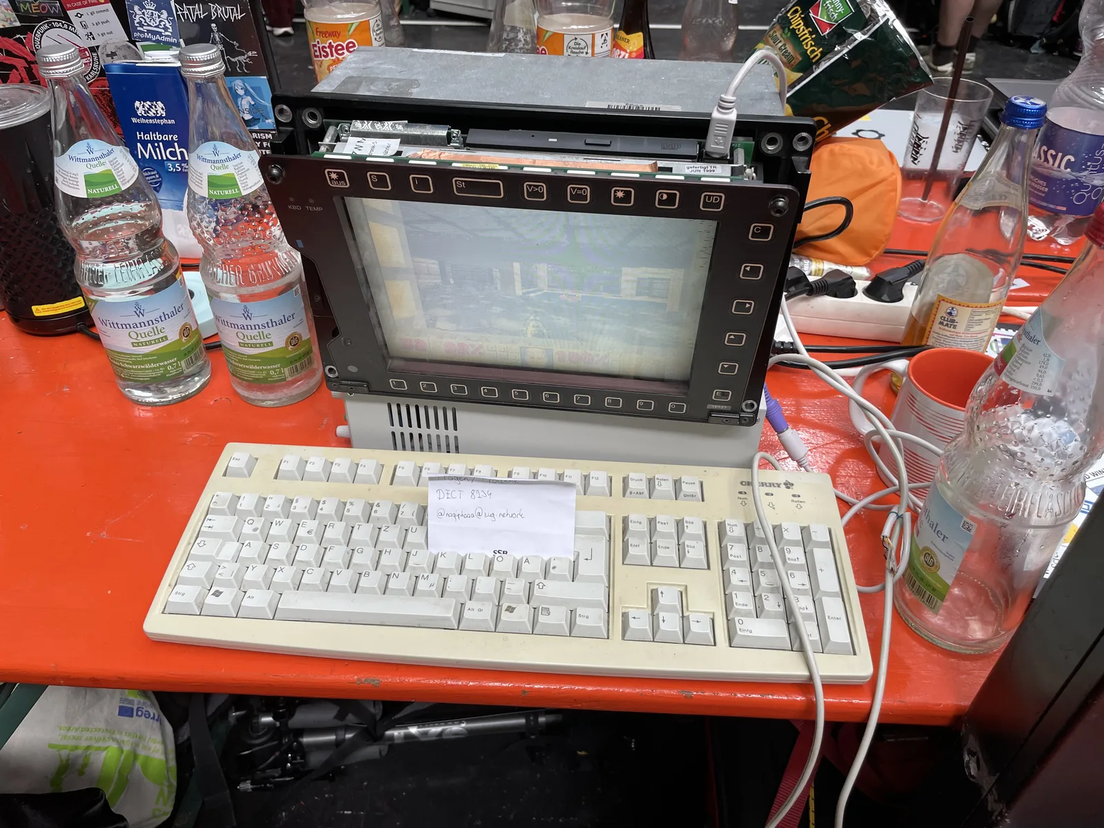 A vintage terminal and keyboard set up on an orange table, surrounded by bottles and a hand-written note.