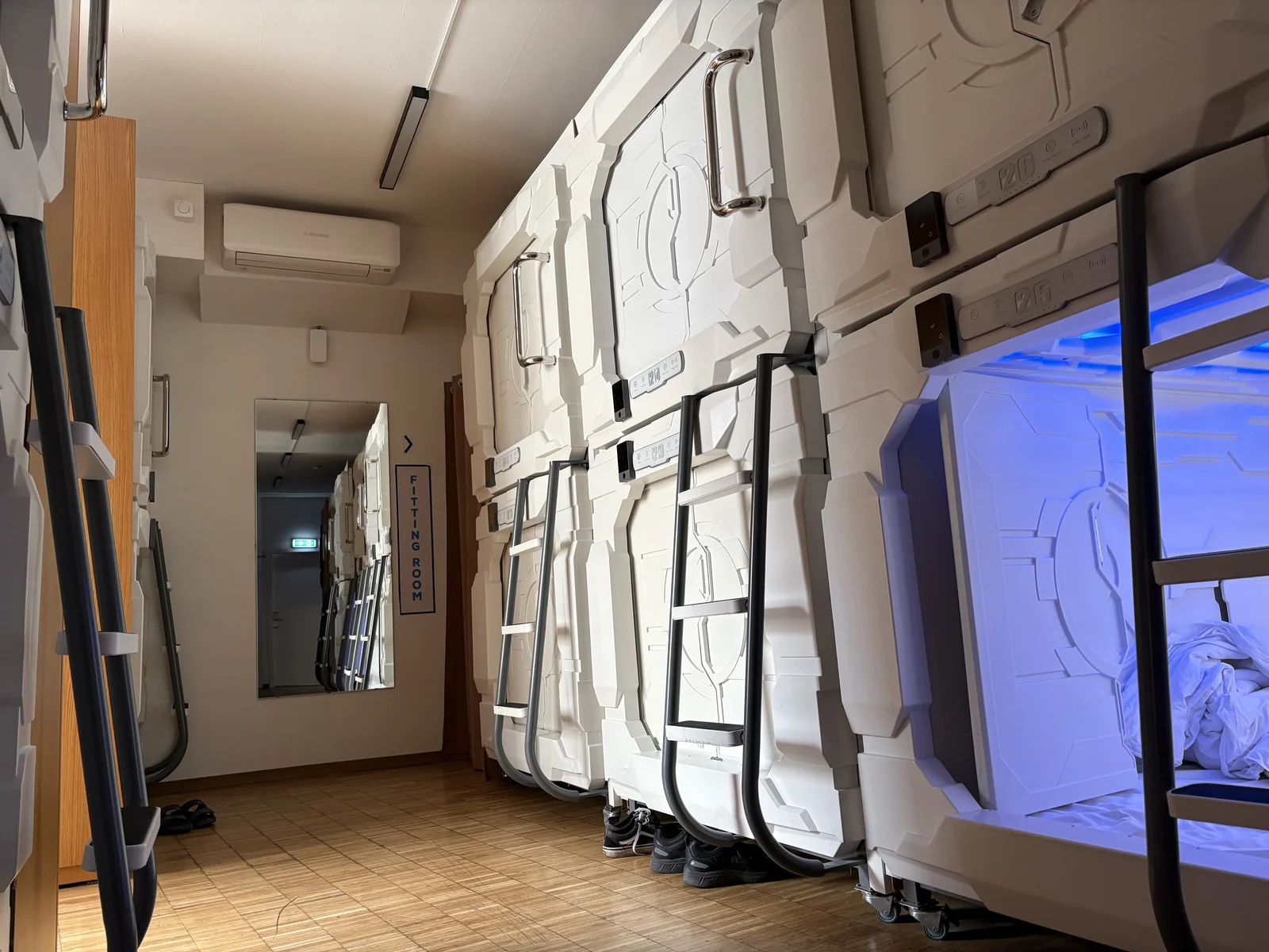 A corridor of stacked white sleeping capsules with metal ladders, one open pod glowing blue, a mirror and a fitting room sign on the wall.