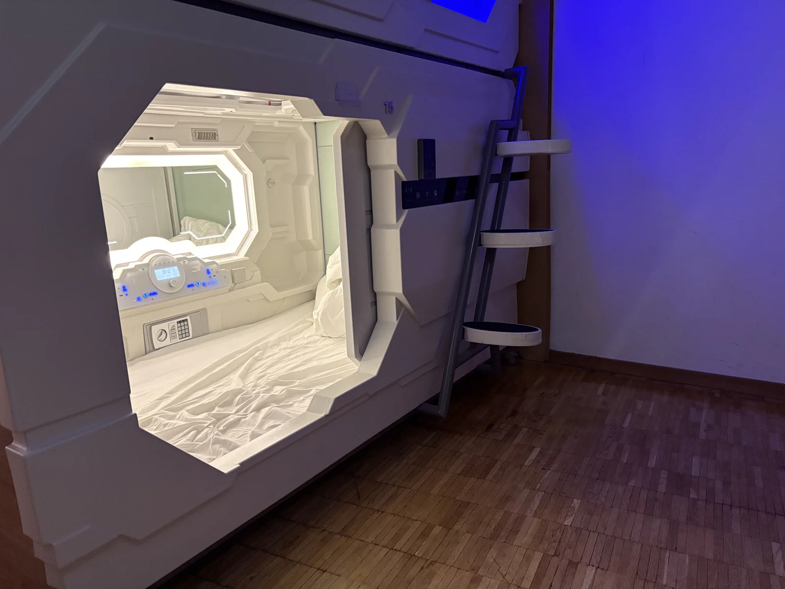 An open sleeping capsule with an octagonal hatch, white bedding, a glowing control panel and a small safe inside, the room bathed in blue light.