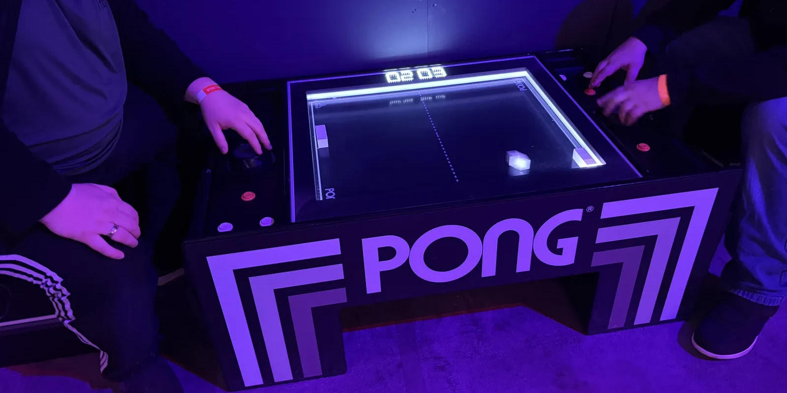 Two players hunched over a glowing Pong coffee table cabinet in violet light.