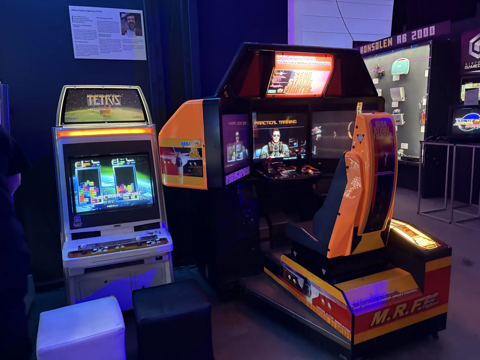 A Tetris arcade cabinet next to a large orange sit-down racing arcade machine.