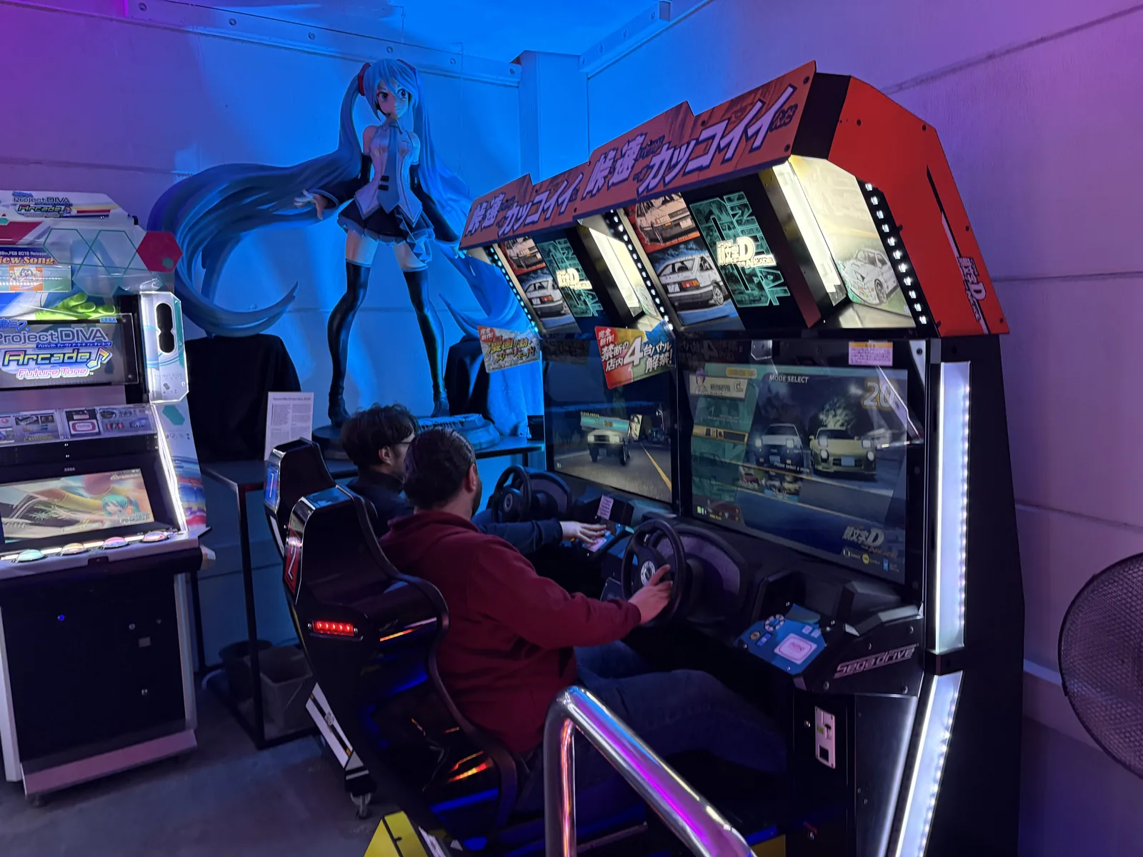 Two people playing a Japanese racing arcade cabinet under blue and purple light, with a large Hatsune Miku figure behind them.