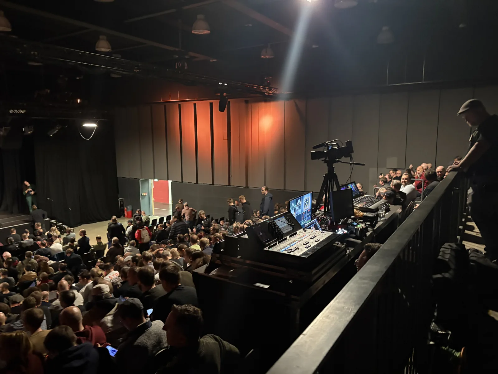 View from the balcony of the AV desk with cameras and a mixing console, over a hall filling with people.