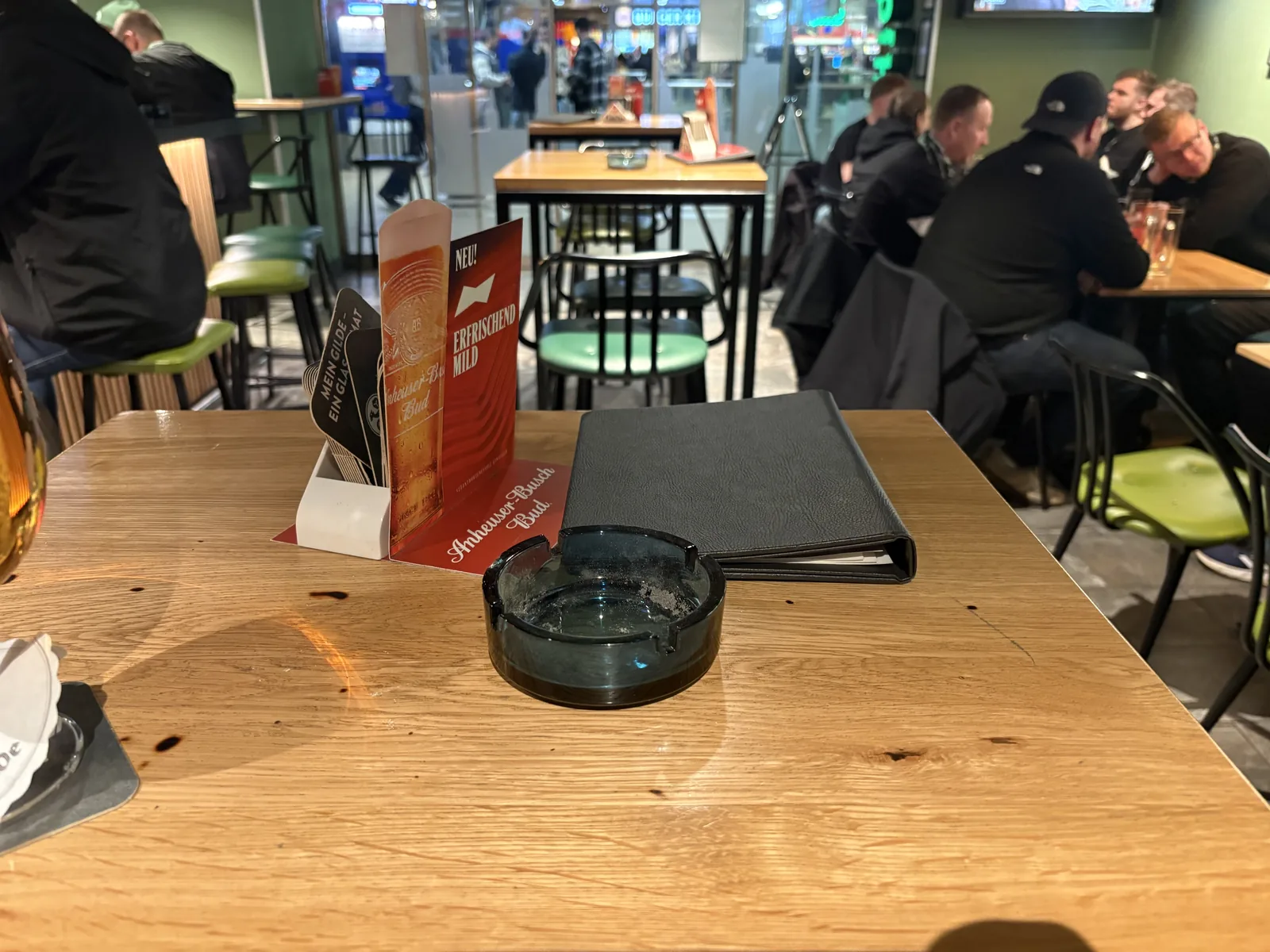 A wooden pub table with a glass ashtray and a menu, with people sitting at tables in the background.