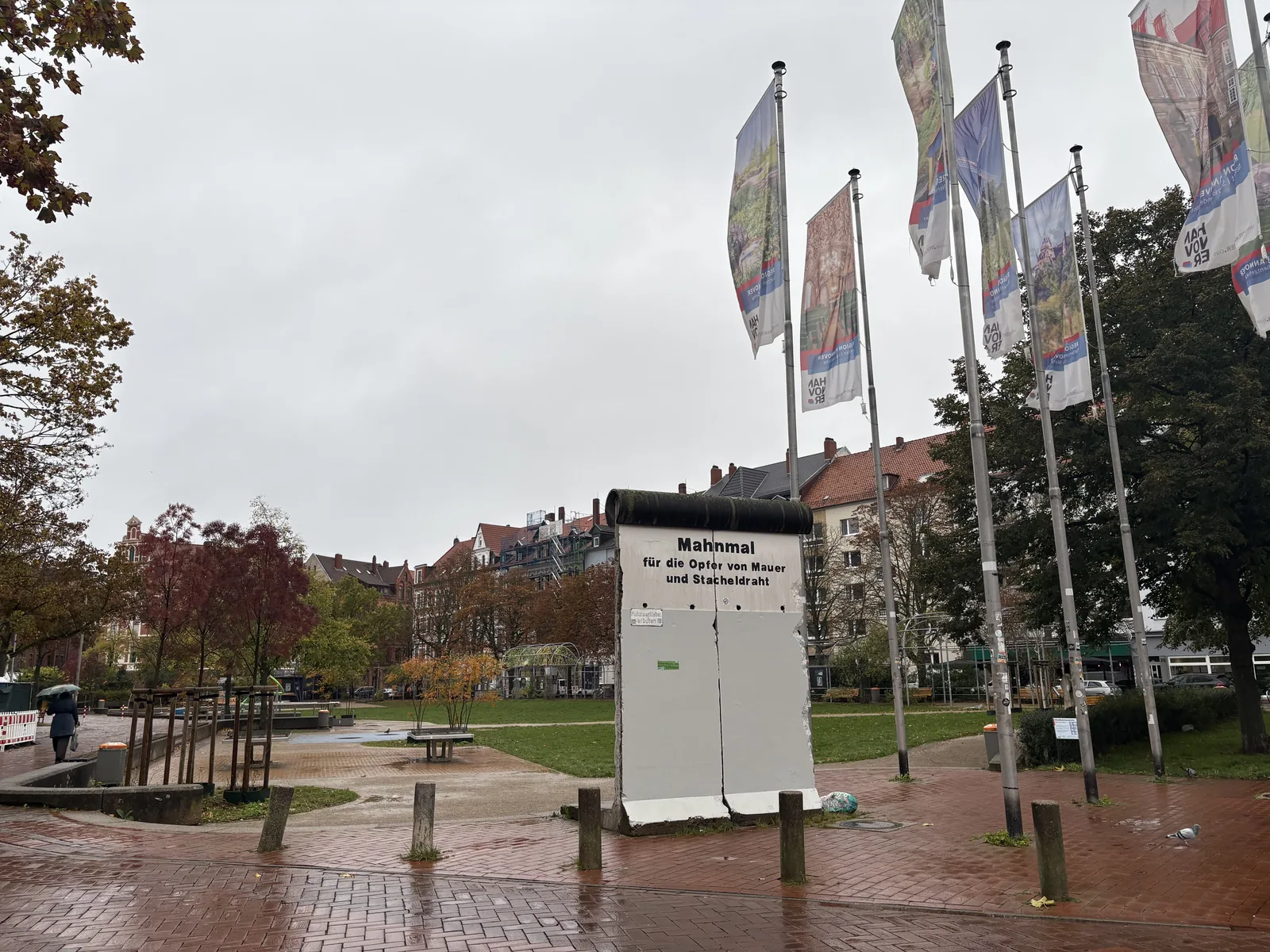 A white segment of the Berlin Wall with the inscription Mahnmal für die Opfer von Mauer und Stacheldraht, next to flagpoles on a rainy square.