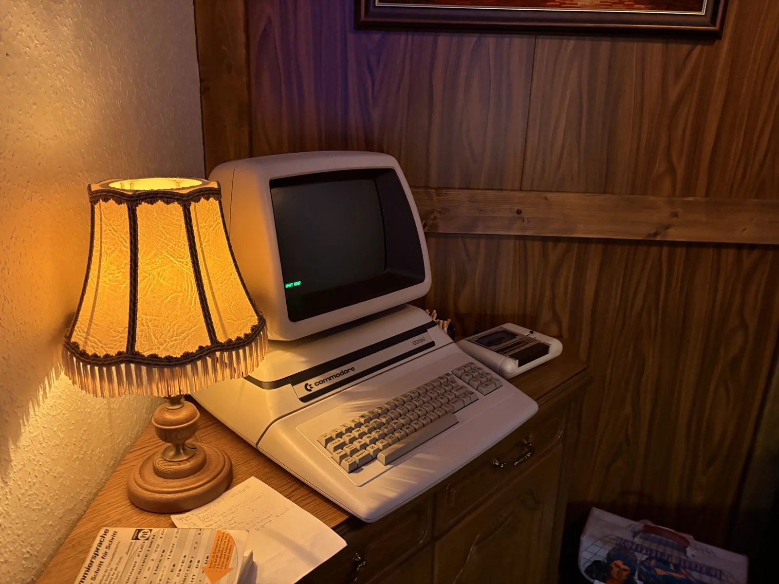 A beige Commodore CBM computer with built-in monitor on a wooden desk next to a fringed table lamp.