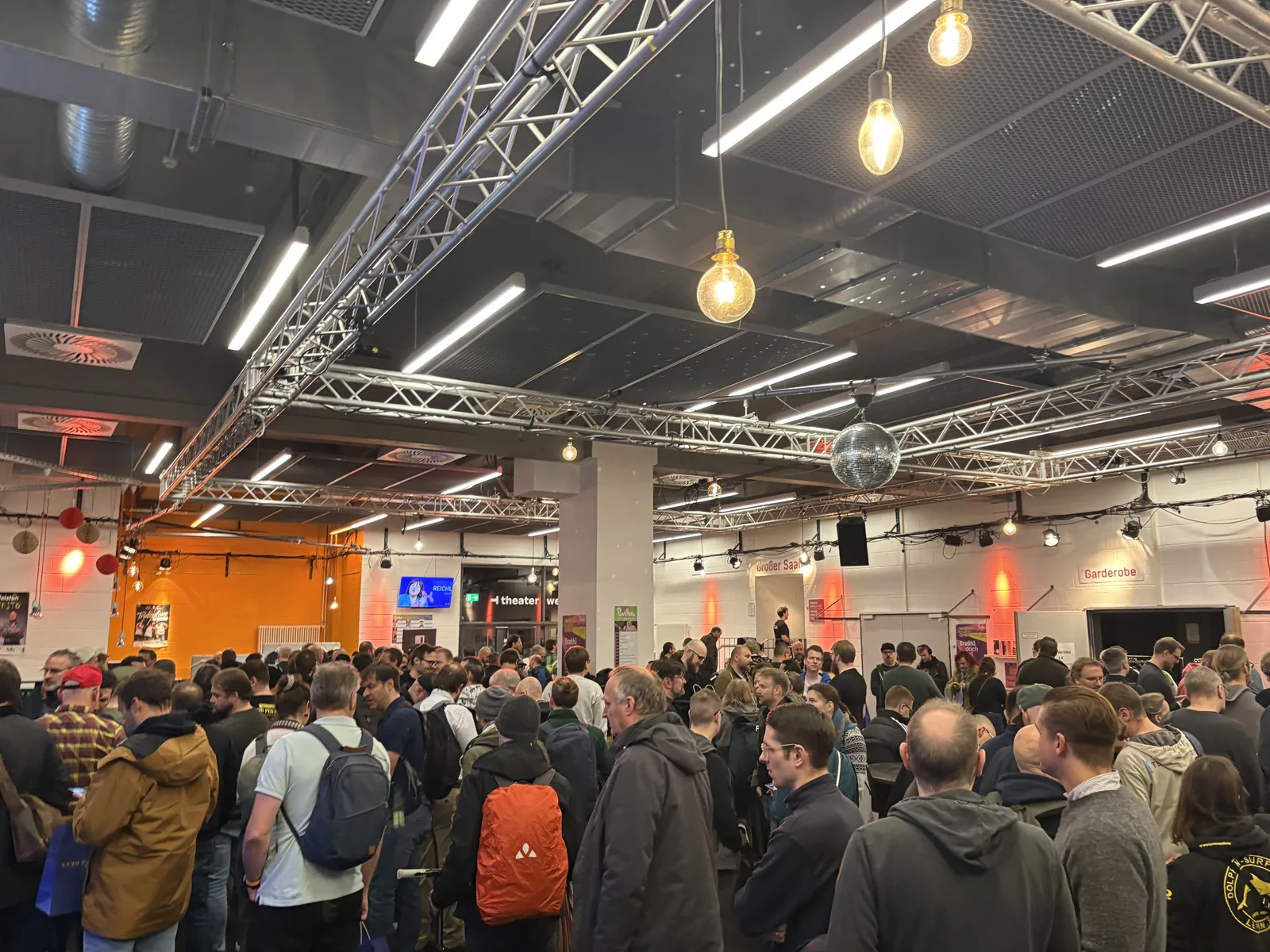 A crowded entrance hall with exposed truss lighting and hanging bulbs, full of convention visitors.