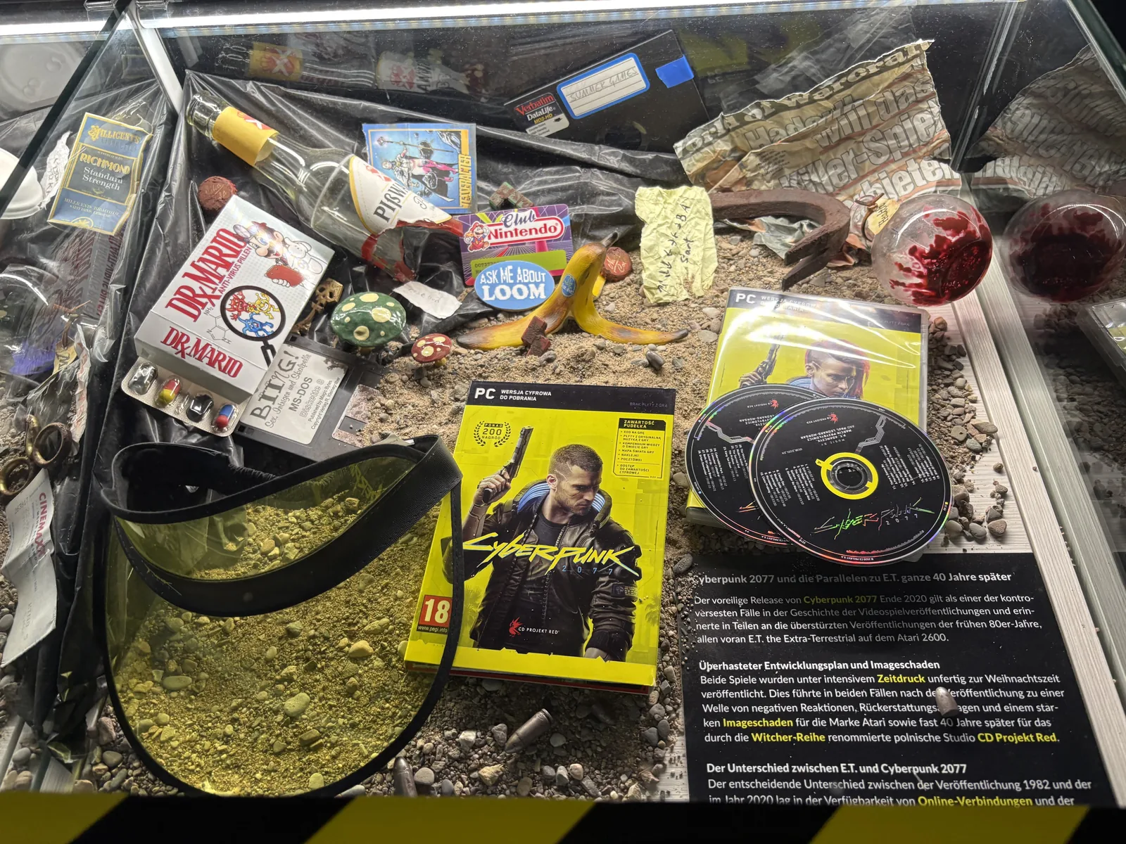 A display case filled with sand and junk, including a Cyberpunk 2077 PC box and discs, with an explanatory text comparing it to E.T.