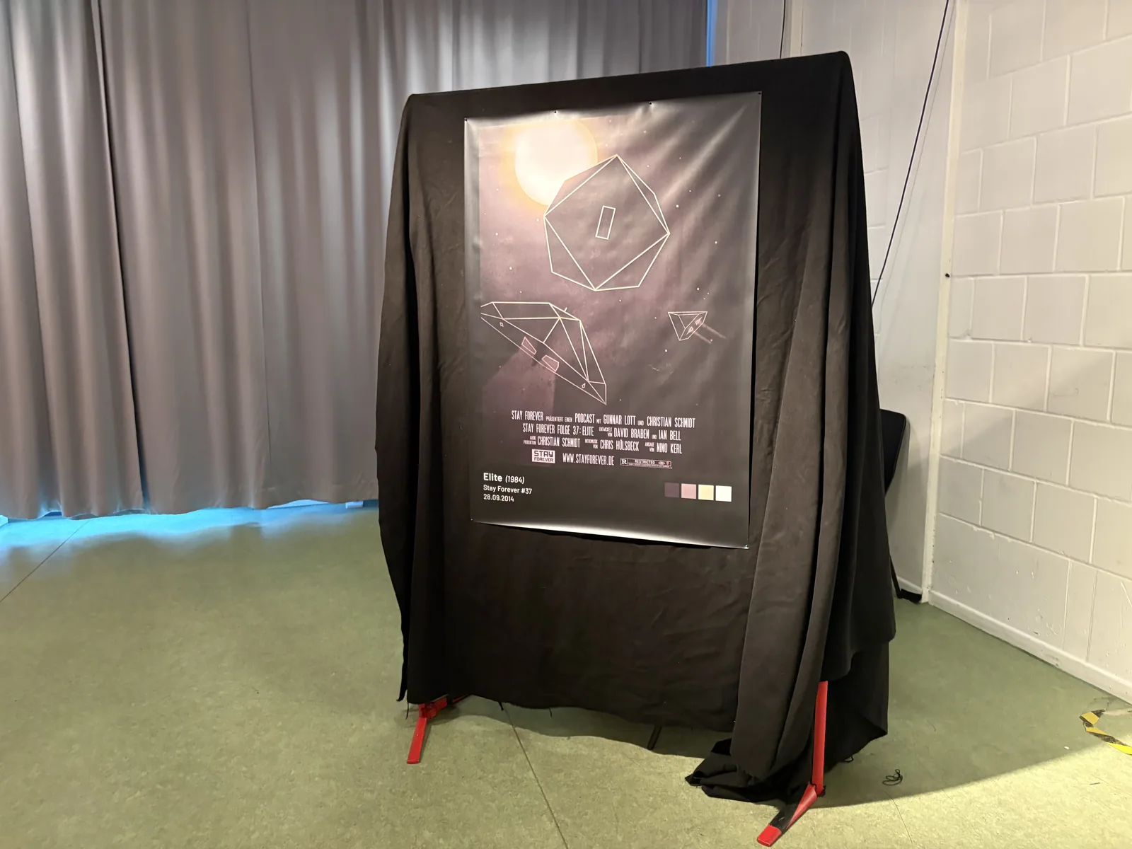 A poster for the Stay Forever episode on Elite, showing a wireframe space station and ships, hanging on a black-draped stand.