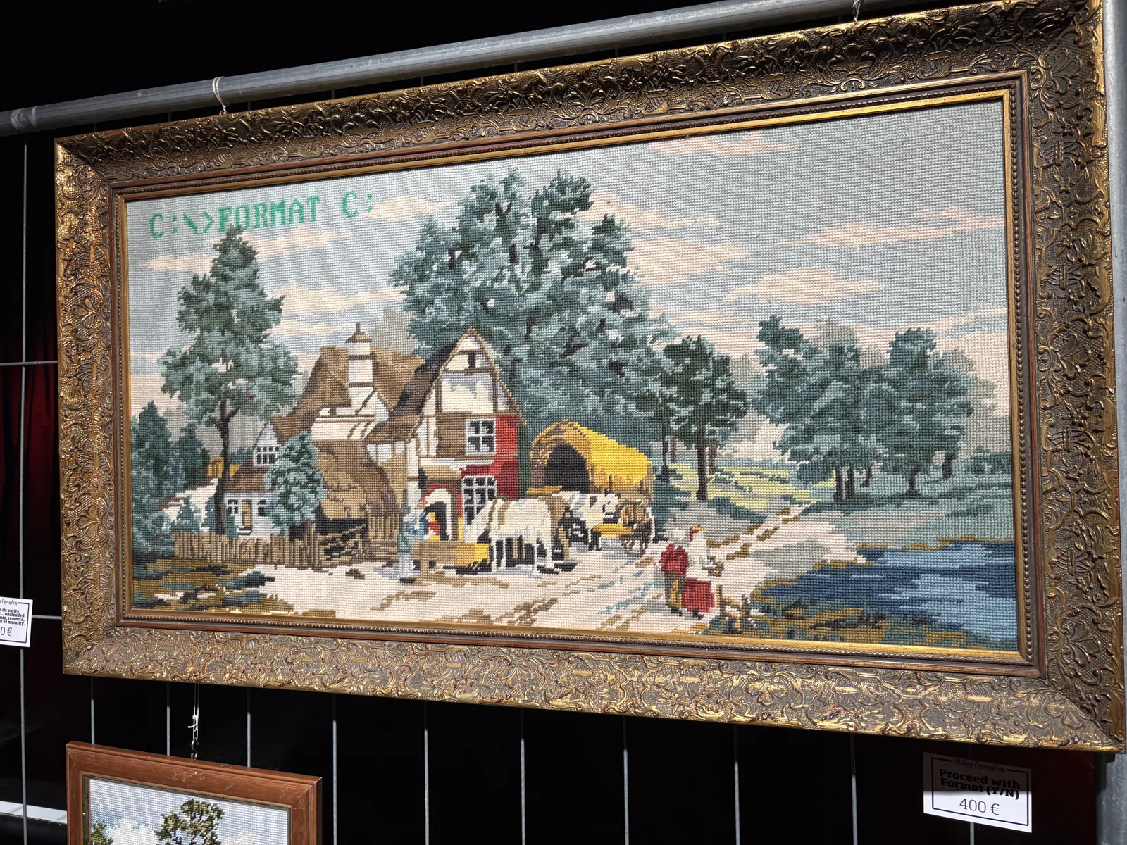A needlepoint tapestry of a rural farmhouse scene in an ornate gilded frame, with the green text C:\>FORMAT C: stitched into the sky.