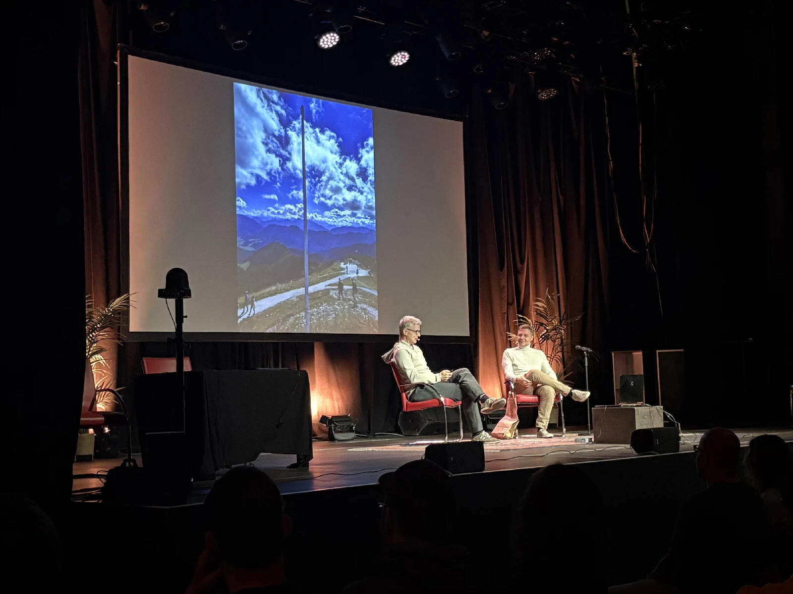 A dark hall with two people seated on a softly lit stage in front of a projection.