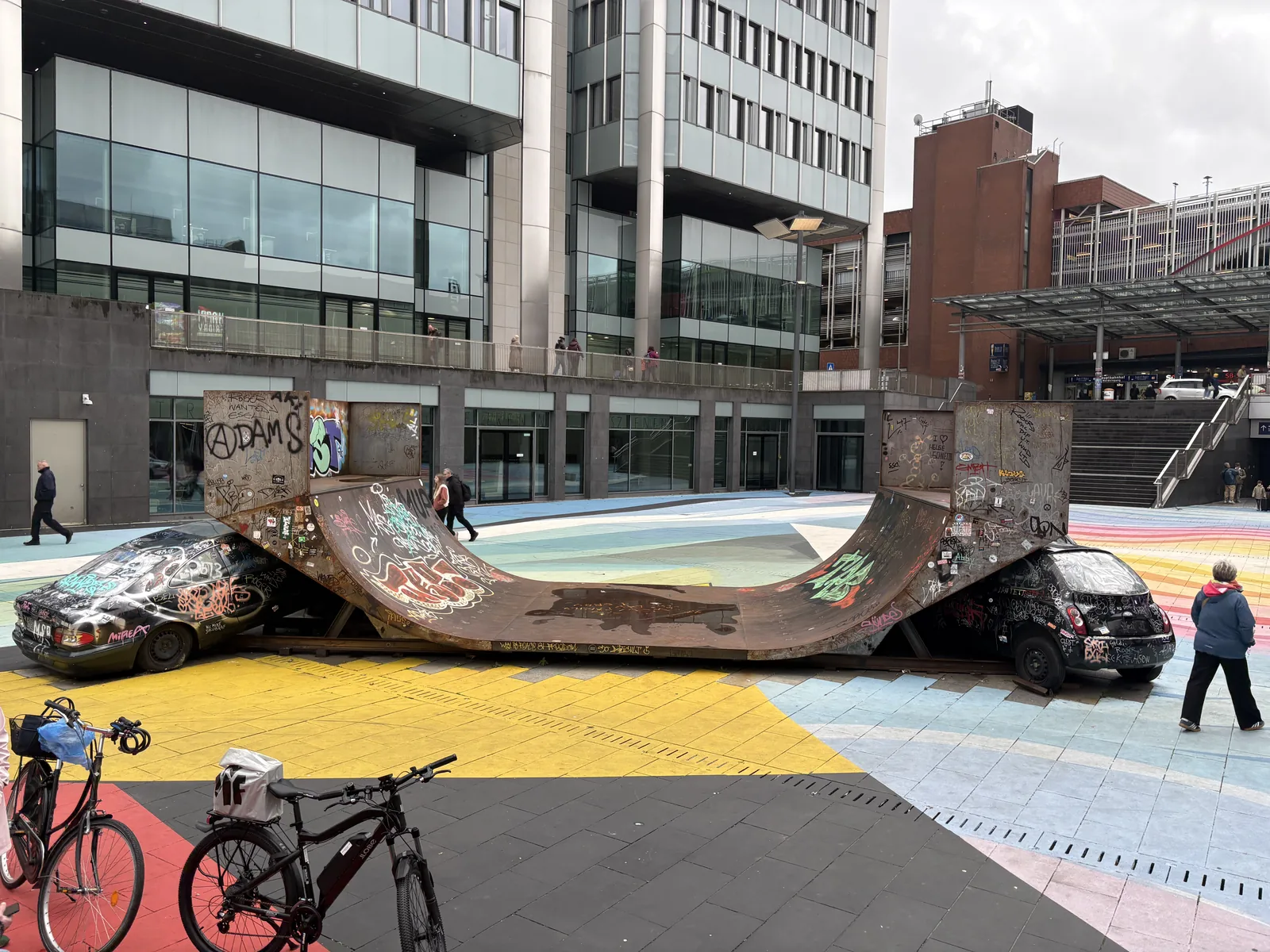 A rusty, graffiti-covered half-pipe art installation resting on two wrecked cars on Raschplatz.