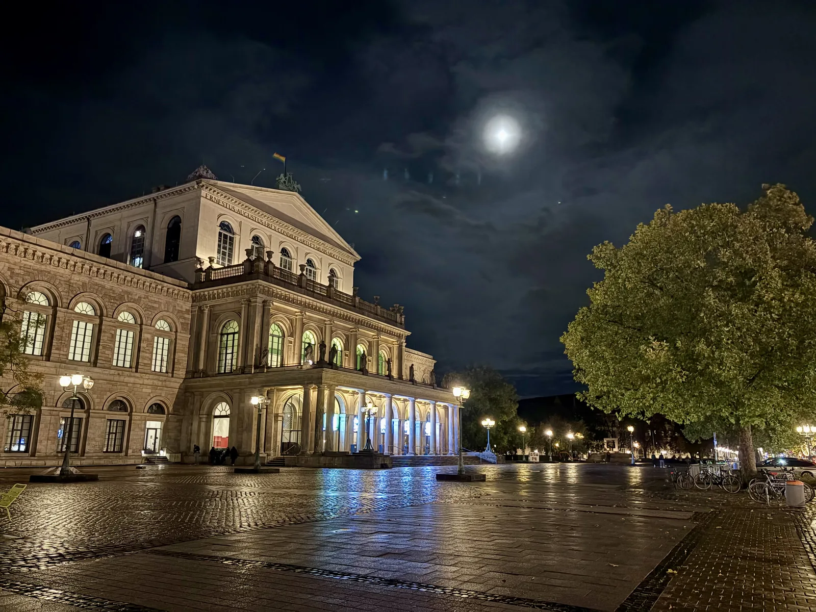 The illuminated Hanover State Opera House at night under a bright moon, with a wet cobblestone square in front.