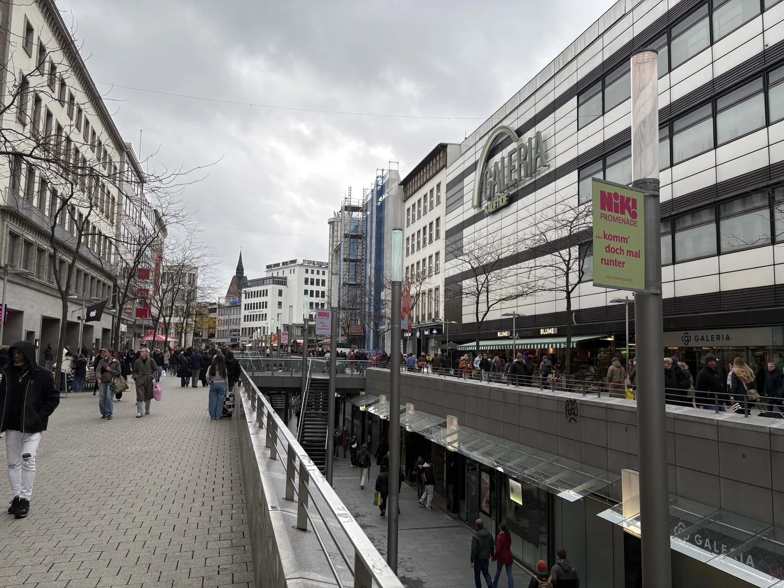 A busy pedestrian shopping street under a cloudy sky, with a sunken passage running alongside.