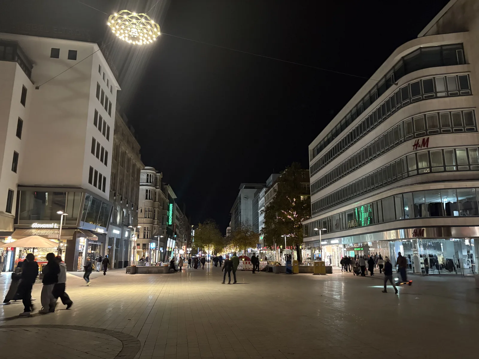A wide pedestrian street at night lit by a large round chandelier-like lamp hanging over it.