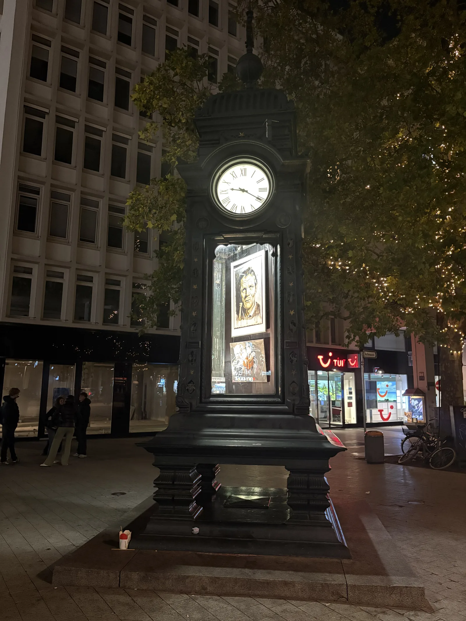 The ornate dark Kröpcke clock column on a square at night, with its clock face lit.