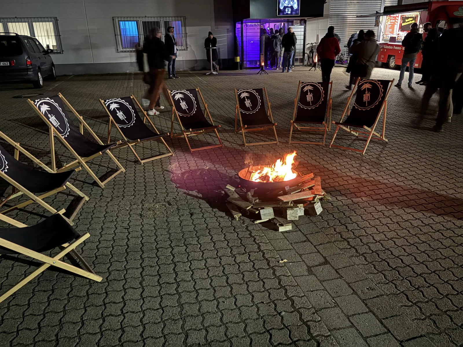 A fire bowl burning on a cobblestone yard at night, surrounded by deck chairs printed with the Stay Forever logo.