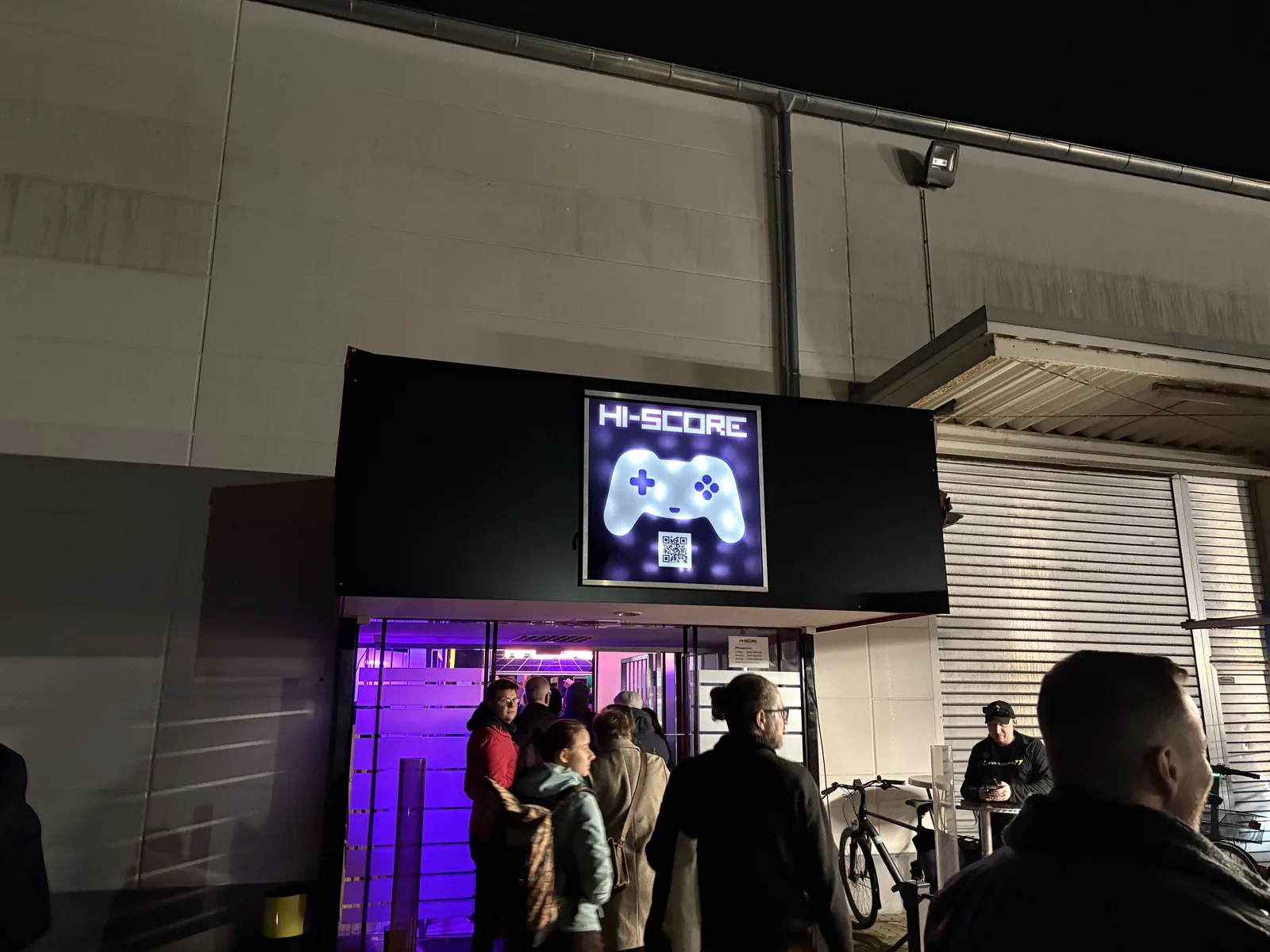 The museum entrance at night with a glowing HI-SCORE game controller sign and people queuing under purple light.