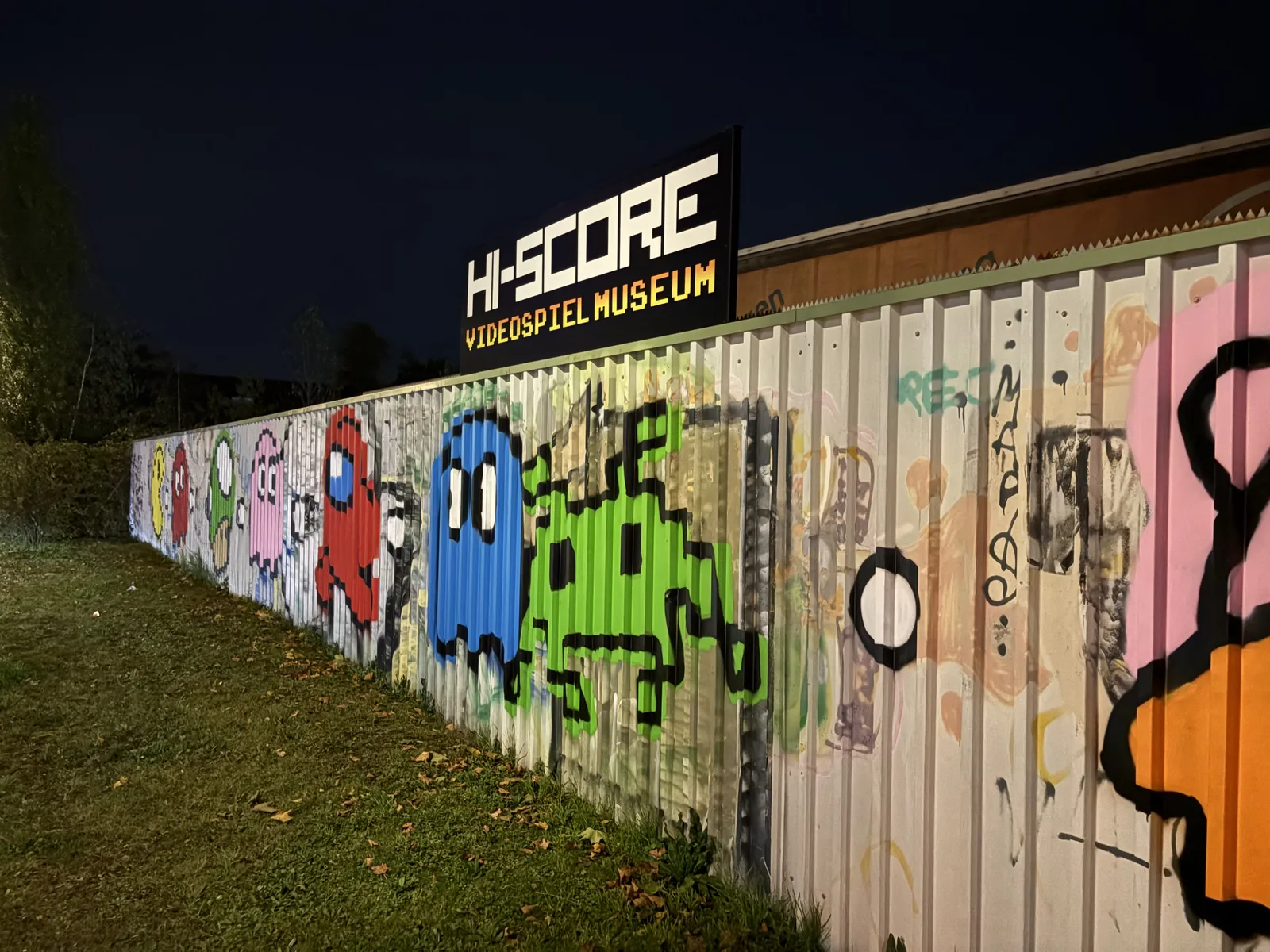 A long corrugated metal wall painted with big graffiti arcade characters and the sign HI-SCORE Videospielmuseum at night.