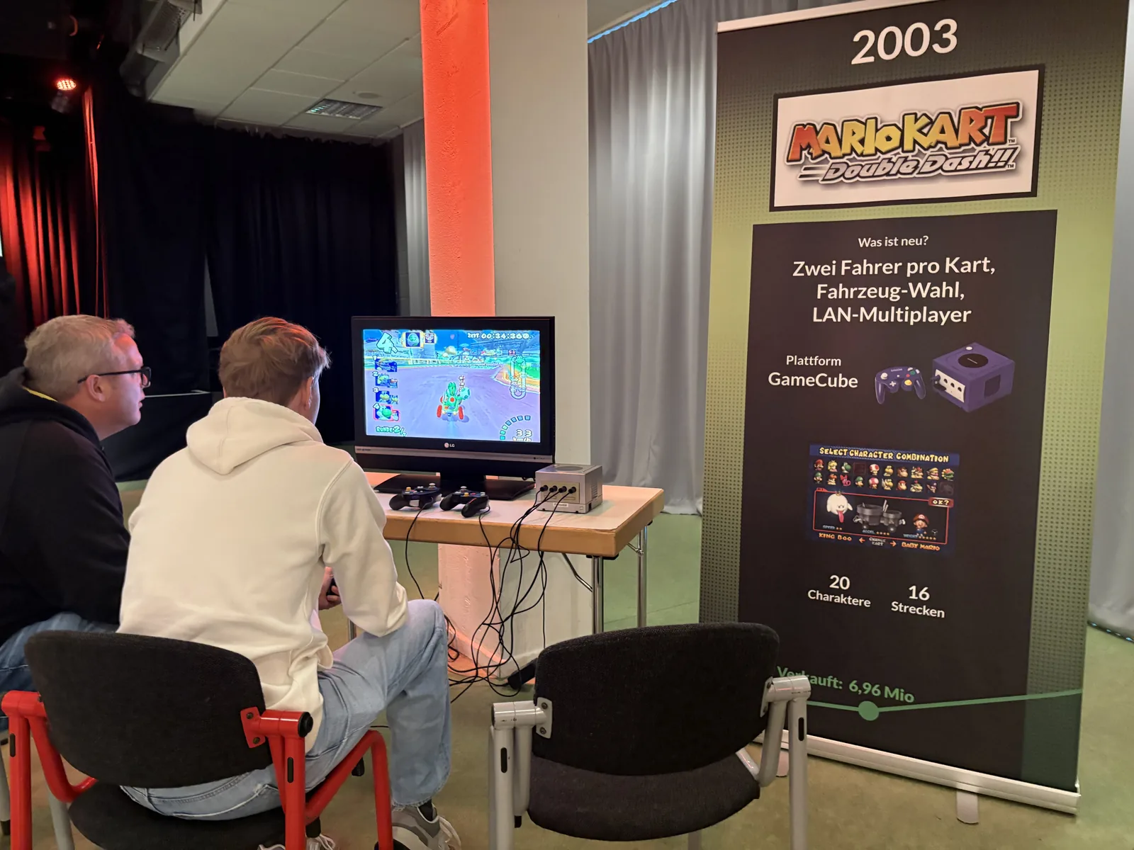 Two people playing Mario Kart Double Dash on a small TV, next to a roll-up banner describing the 2003 game.