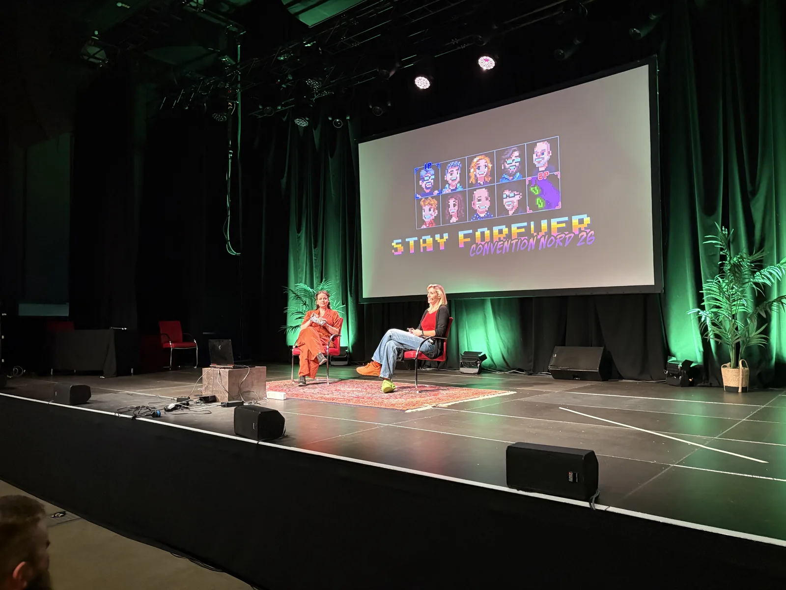 Two women seated on a stage with green lighting and plants, in front of a screen with the Stay Forever logo.