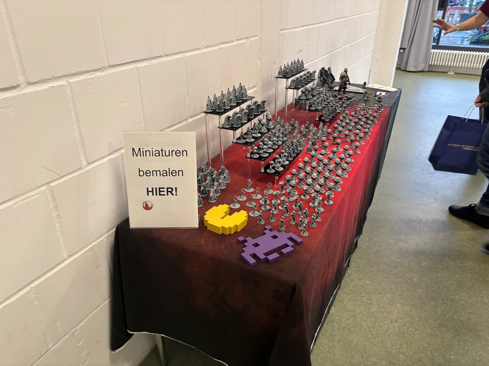 A table of grey unpainted miniatures with a yellow Pac-Man and a purple Space Invader, next to a sign reading Miniaturen bemalen hier.