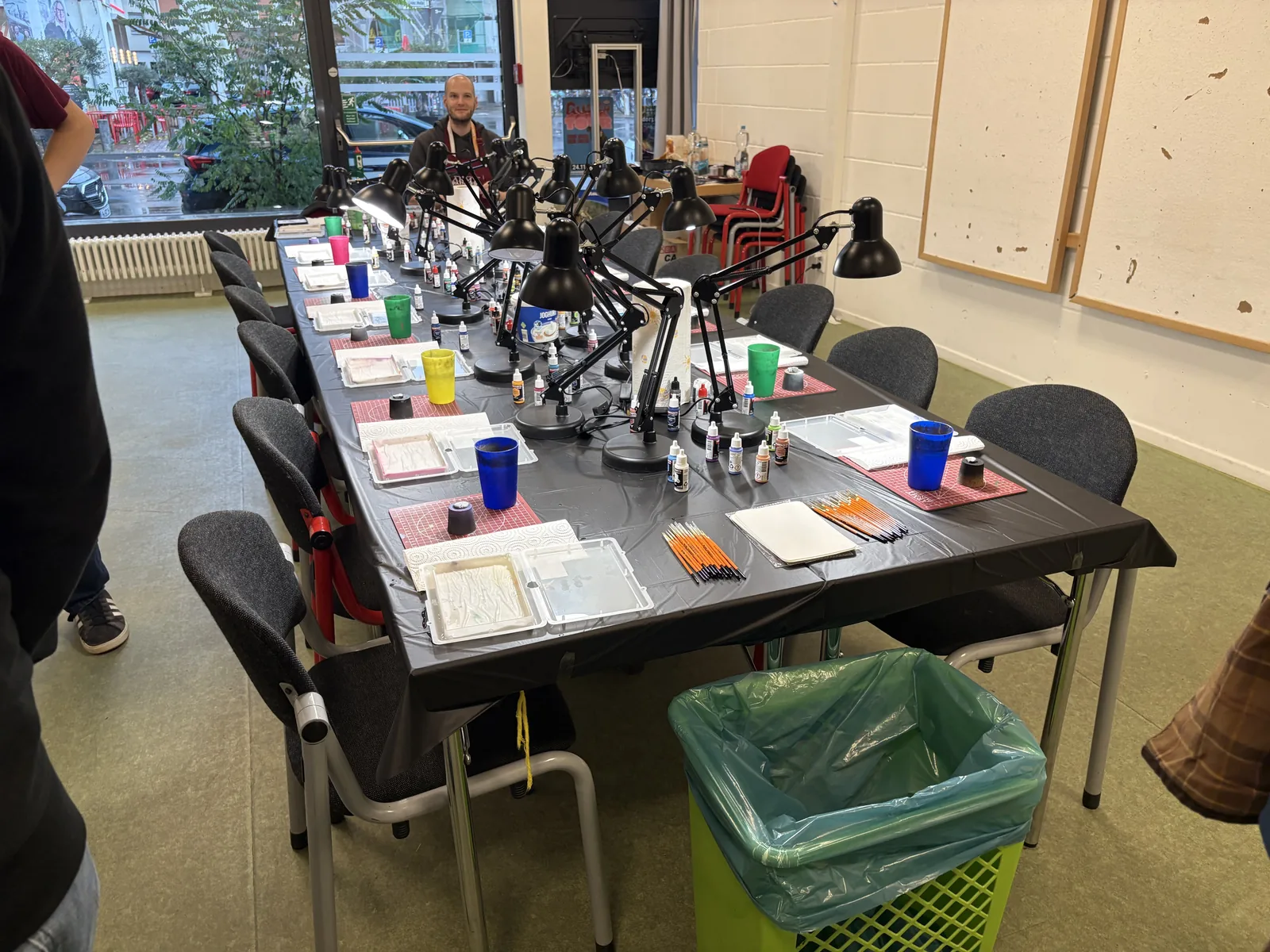 A long, empty table set with desk lamps, paint pots, brushes, and water cups.