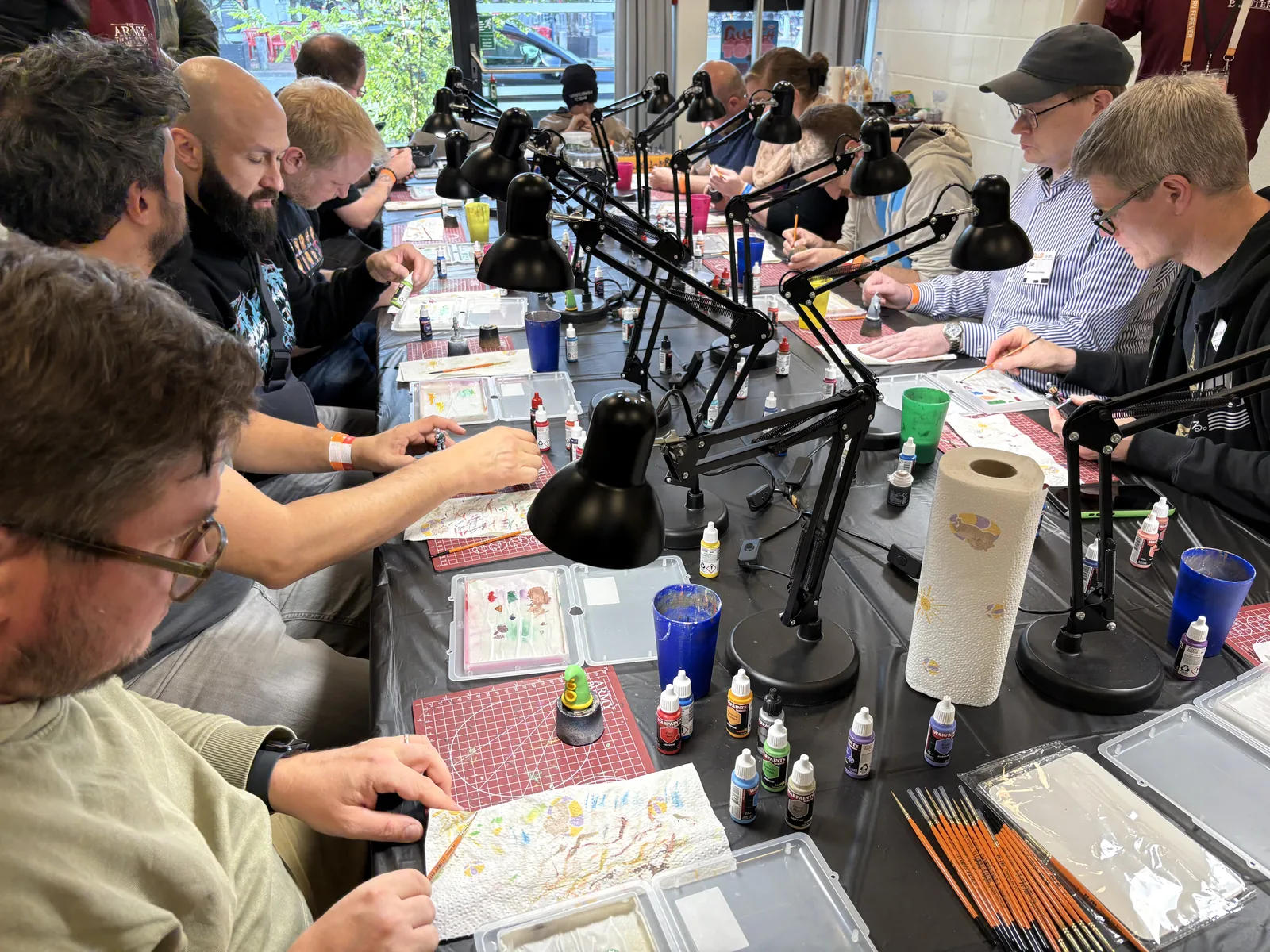 Convention visitors seated along a long table, concentrating on painting miniatures under desk lamps.