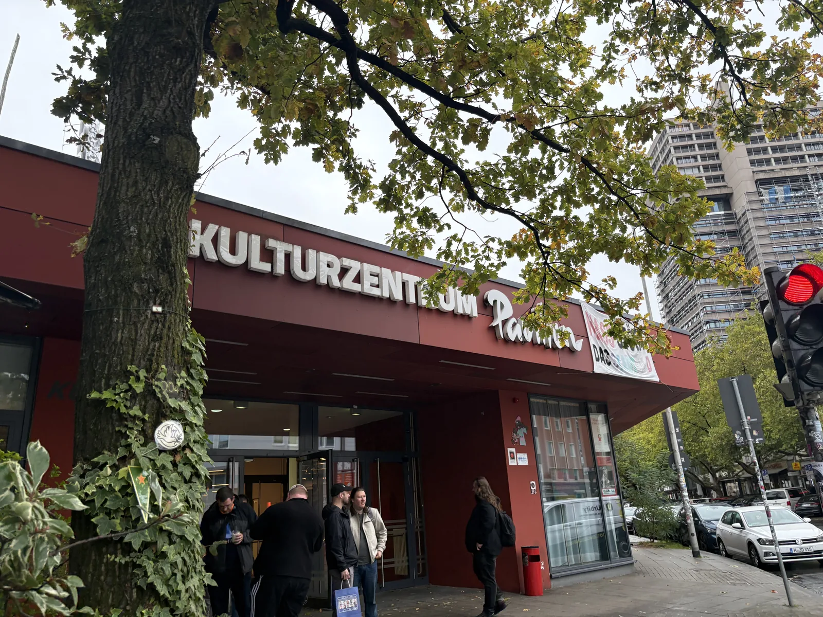 The red entrance front of the Kulturzentrum Pavillon behind a large tree, with people arriving.