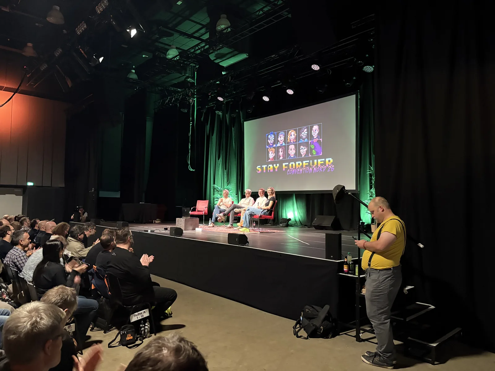 An audience member at a microphone asking a question of the team seated on the green-lit stage.