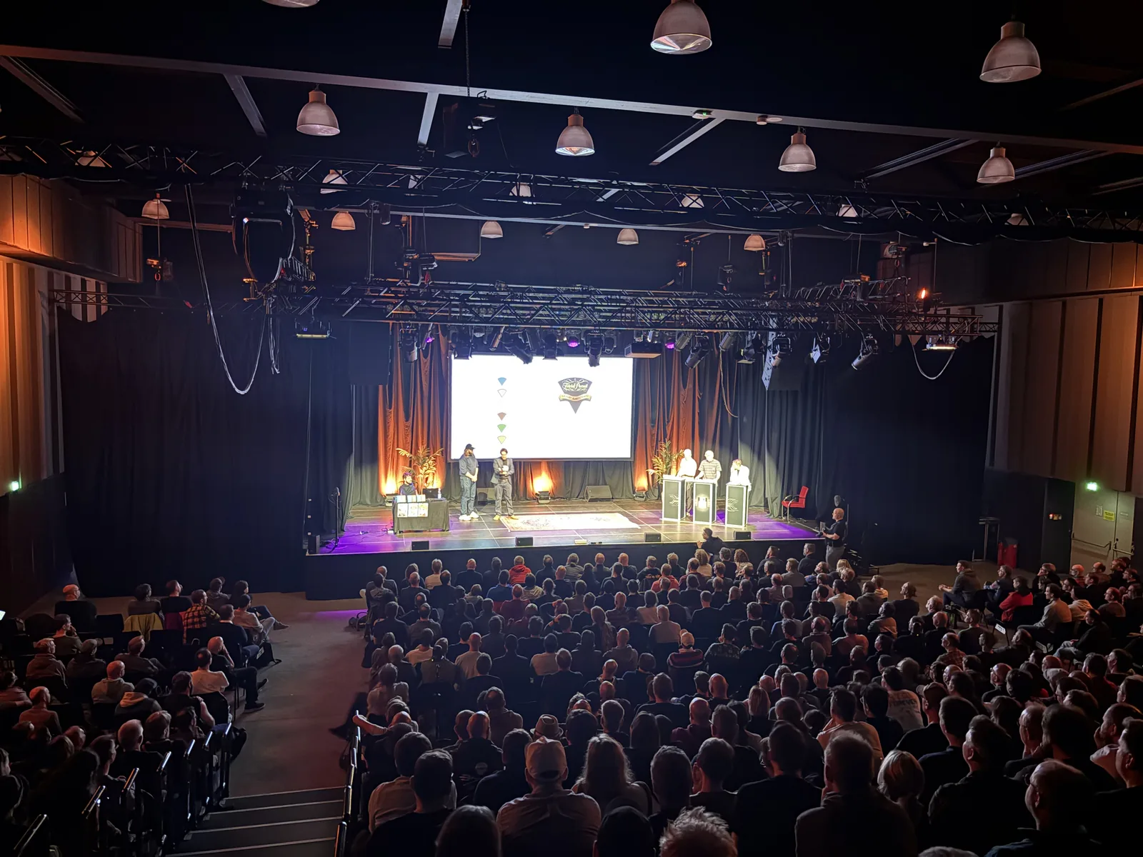 A full concert hall watching the evening quiz on a brightly lit stage with candidate podiums and a large screen.