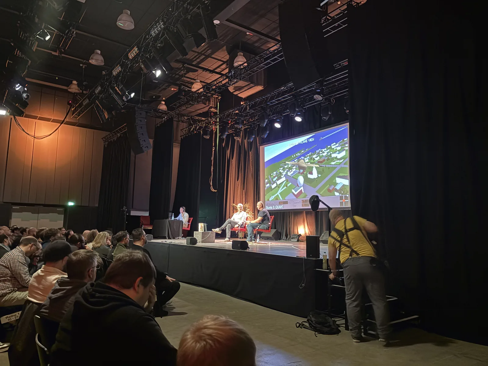 The stage seen over the audience's heads, with three hosts in armchairs and a projection of a 3D flight scene from Stunt Island.