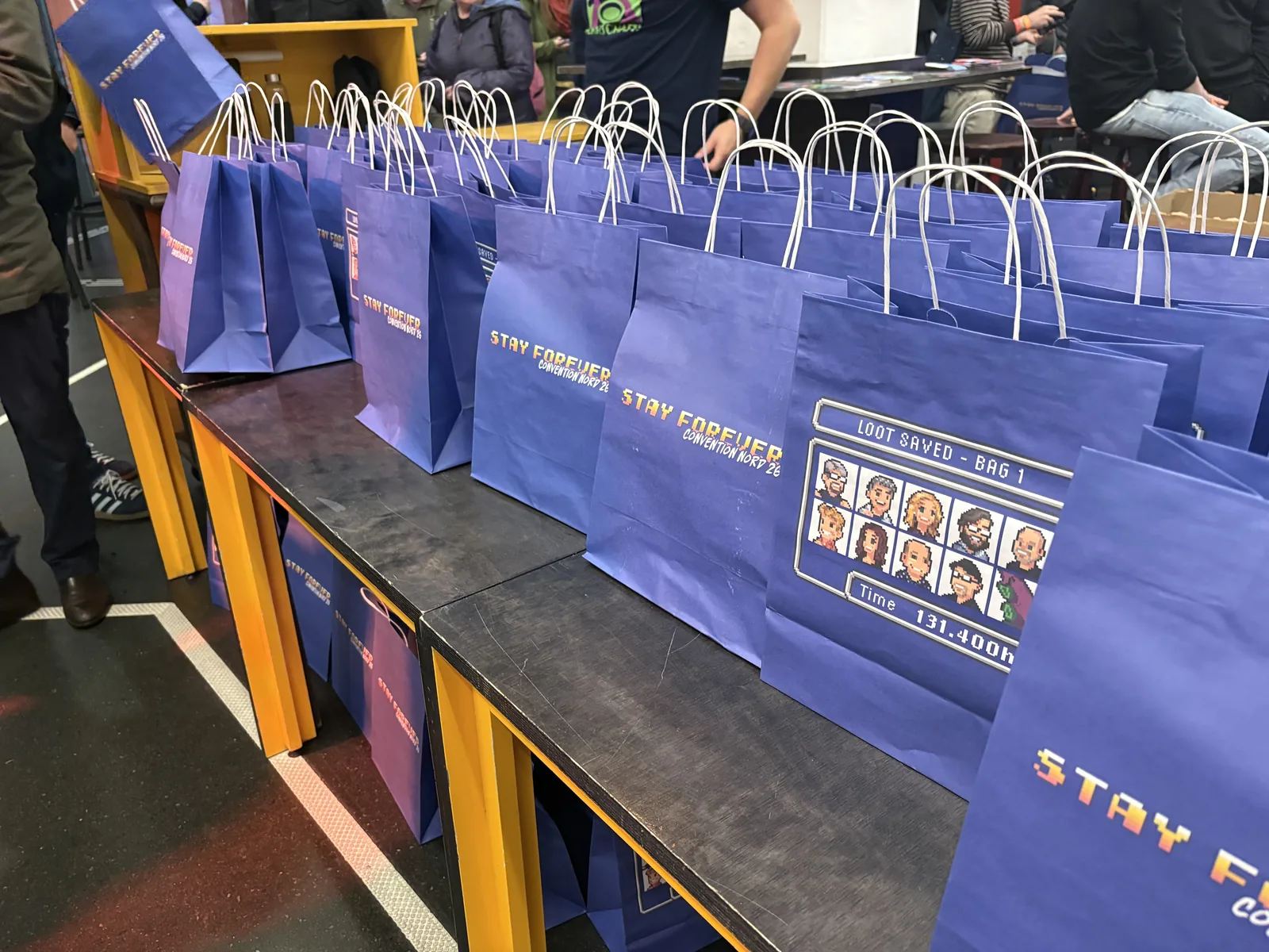 Rows of blue Stay Forever paper bags printed with pixel art portraits of the podcast team.